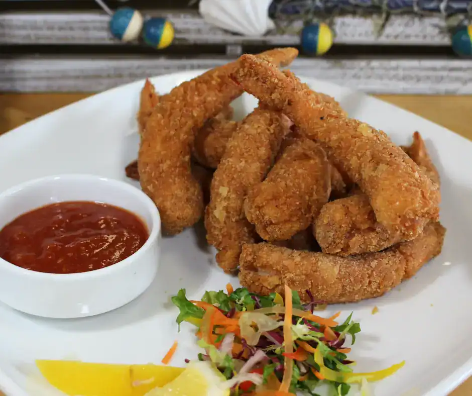 Crispy Island Shrimp Platter with Dipping Sauce Breaded fried shrimp on a white plate with red dipping sauce, lemon wedges, and mixed greens garnish.