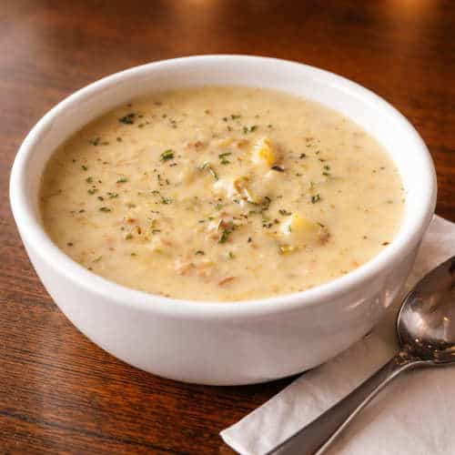 Creamy Shrimp and Corn Chowder – The Original Island Shrimp House Close-up of a white bowl of creamy New England clam chowder with potato chunks and herbs, served on a wooden table with a spoon on a napkin.